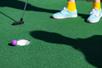 Close up of a purple golf ball sitting on the edge of the hole with a person wearing yellow socks and blue shoes in the background.