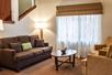 A sitting area with a brown sofa, two light brown cushioned chairs, oval coffee table, side table, lamp, and curtained window.