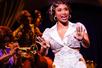 A woman with short dark hair in an ornate white dress singing with her hands up with other women playing jazz instruments in the background.