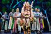 A man dressed as a peasant carrying a pack of things on his back with six knights behind him with their hands together on stage in the musical Spamalot.