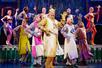 The cast of Spamalot lined up in rows dancing with their arms up with the king dressed in gold smiling in the front.