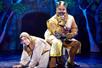 A king in a bright gold costume sitting on a peasant wearing rags on stage in the musical Spamalot.