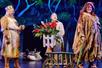 A king wearing gold on the left, a large woman wearing rags on the right, and a peasant man in the middle holding a box of flowers in the musical Spamalot.