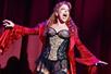 A woman with red hair wearing black lacy lingerie and a bright red velvet overcoat with bell sleeve sining on stage in Spamalot.