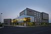 Street view of the SpringHill Suites Denver Downtown with the sun setting behind it and some lights visible through the large glass windows on the corner of the building.