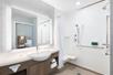 A modern white bathroom with a large vanity with a sink in the middle and a lit mirror over it, and an accessible shower with a bench and grab bars.