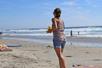 A woman in a tank top and shorts playing bean bag toss on the beach with the ocean in the background.