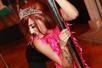 A woman with red hair wearing a bright pink feather boa and a crown while leaned over holding onto a silver stripper pole.