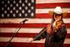 A woman with long brown hair wearing a ivory cowboy hat and playing the fiddle with an American flag behind her.