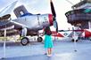 A little girl in a bright blue dress standing at a guard rope looking at the older gray plane on the other side of it at The Intrepid Sea, Air & Space Museum.