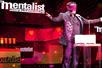 A man in a black suit wearing a red blind fold standing on stage behind a podium with his arms out at The Mentalist in Las Vegas.