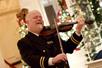 A bald man dressed in a black uniform playing a dark brown violin with Christmas trees and decorations behind him.