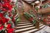 A grand wooden staircase leading up with garland, lights, and bows lining the railing and Christmas trees at the bottom.