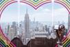 A tower viewer in front of a rainbow heart on the glass at the Top of the Rock Observation Deck with New York City in the background.