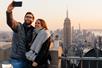 A couple in winter clothes standing taking a selfie with the Empire State Building in the background at sunset.