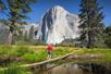 A hiker in a red coat balancing on a log over a river on a sunny day in Yosemite National Park.