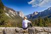 A hiker in a white shirt sitting on a rock wall over looking Yosemite National Park on a sunny day with clouds in the sky.