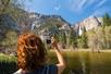 A hiker with bright red hair taking a picture of a mountain with a lake in front of it on a sunny day in Yosemite National Park.