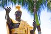 The King Kamehameha Statue, a man dressed in gold holding a spear with his other hand out with palm trees behind it.