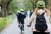 Tourist wearing neon green helmets and riding bikes on a paved path in a park with lots of trees, and people walking to the right of them.
