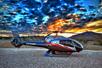 A silver, black and red Maverick Airbus helicopter on the ground with and bright blue and orange cloudy sky overhead.