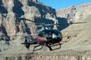 A silver, black, and red Maverick helicopter flying through a rocky canyon on a sunny day.