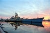 A large ship in the water at the Battleship New Jersey Museum and Memorial, seen against a backdrop of the sky and water, with reflections in the water. The scene captures the ship during either sunrise or sunset.
