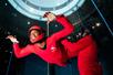 Two men in red jump suits in a flight tunnel, one of them floating in the air and the other holding onto his suit guiding him.