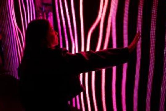 A woman in a jacket in a dark room with her arm out touch stripes of different shades of pink lights in front of her.