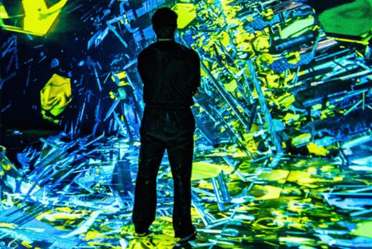 The silhouette of a man facing away from the camera looking of bright green and blue art on the floor, walls, and ceiling.