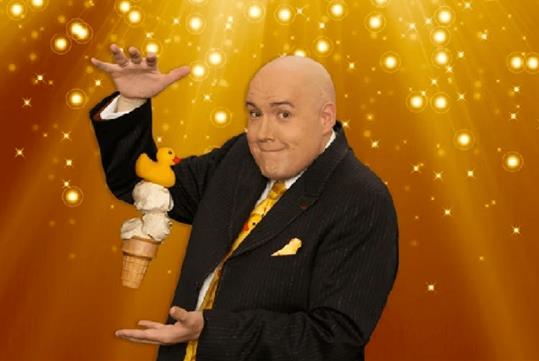 Adam London in a black suit maneuvering an ice cream with the rubber duck on top that seems to be levitating between his hands.
