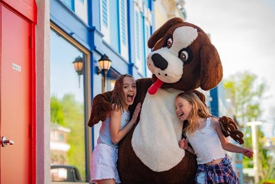 Two young girls laughing and hugging the Bernie Bernard mascot with colorful buildings on the background at Adventureland.