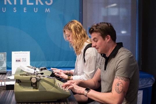 A man and woman sitting next to each other typing on old green typewriters  at the Story of the Day exhibit at the American Writers Museum.