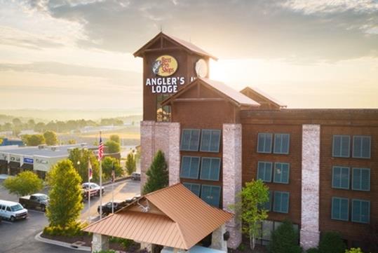 The front exterior of the Angler's Lodge Hollister with the sun shining brightly behind it and cars in the parking lot to the left.