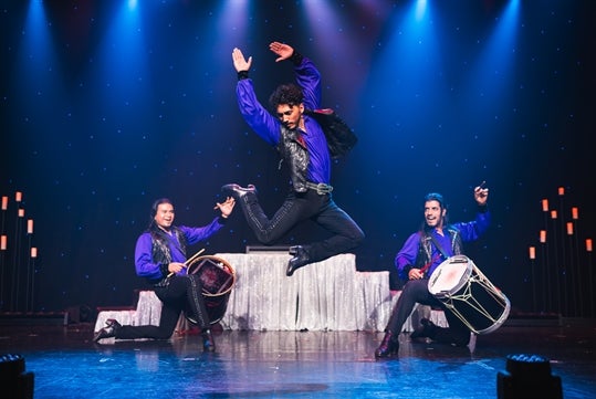 Two men are playing their drums hyping up the dancer as he jumps into mid air performance all wearing purple and black costumes.