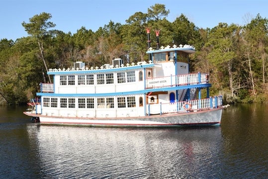 A white, red and blue double-decker river boat with the "Barefoot Queen" in the water with a line a trees behind it on a sunny day.