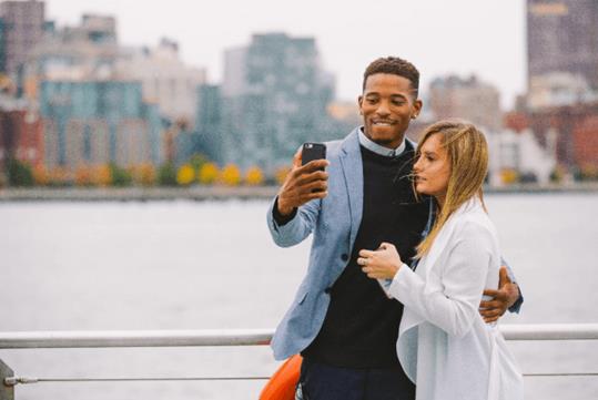 A man and woman standing near the railing of a cruise boat taking a selfie with water and the city in the background on the Bateaux New York Premier Brunch Cruise.