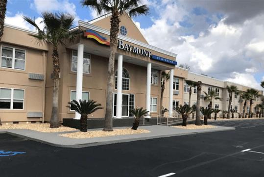 The front entrance of the Baymont by Wyndham with several palm trees in the landscaping in front of it and a blue cloudy sky over it.