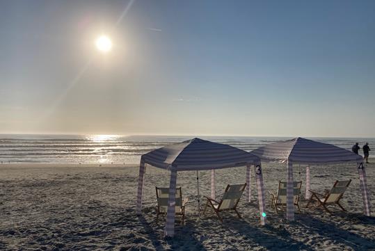 Two striped cabana beach tents with two chairs sitting under each of them with the sun shining over the water in front of them.