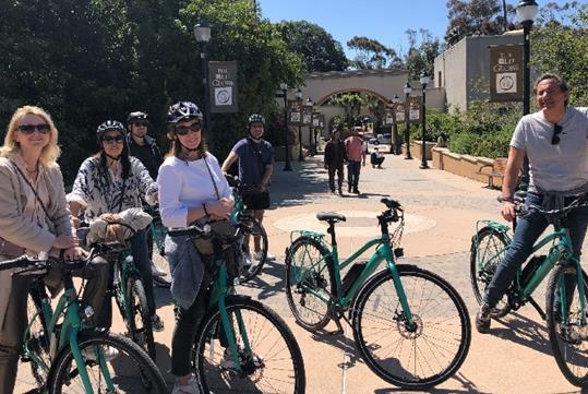 A group of tourists on green bikes stopped on a brick walkway with other people walking behind them on a sunny day in San Diego.