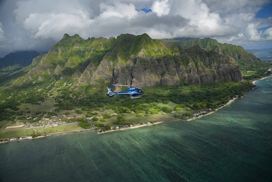 A helicopter flying over the ocean with the huge mountains of Oahu behind it and a small town along the coastline on the Blue Skies of Oahu Helicopter Tour in Hawaii USA.