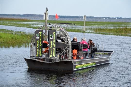 A black air boat full of people in the water with marsh grass in front of them and trees in the distance at Boggy Creek Airboat Adventures.
