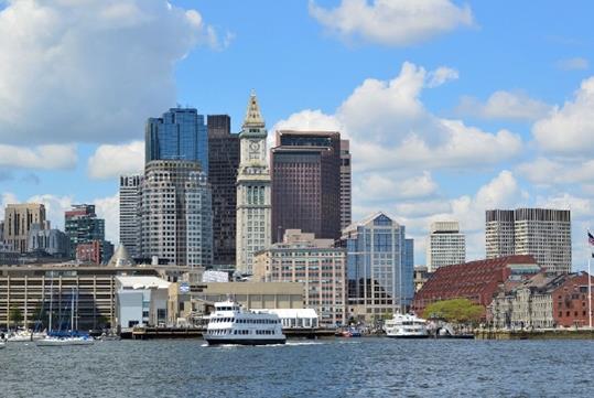 Wide shot of the Boston Harbor with a large white cruise boat in the water with the city behind it and a blue sky full of clouds overhead.
