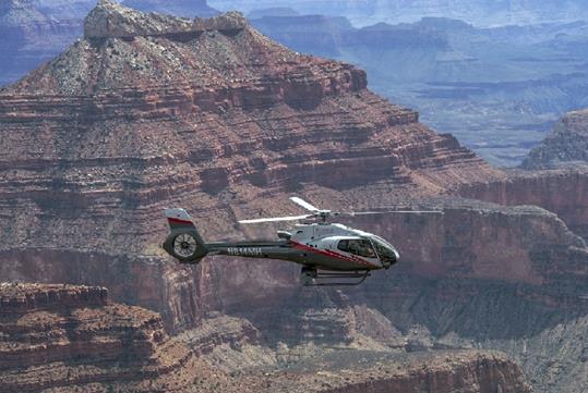 A Maverick Airbus helicopter flying by Point Imperial and Marble Canyon with the rocky formations in the background on a sunny day.