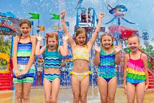 A group of five young girls in their swim suits laughing while water plashes down on them and a water jungle gym behind them.