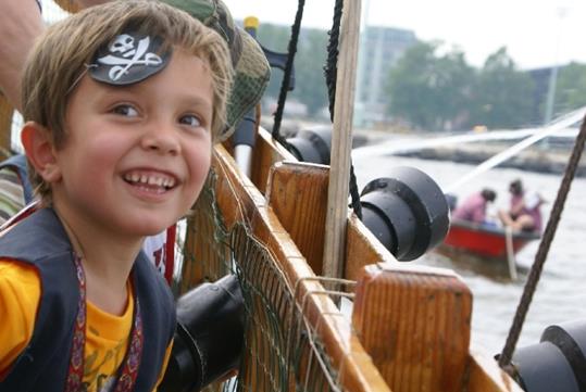 A smiling little boy in a black vest with a pirate patch on his forehead near the railing of the Sea Gypsy boat.