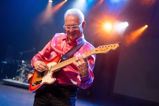 A man in a shiny pink shirt playing with his guitar on the stage with a drum set in the background.