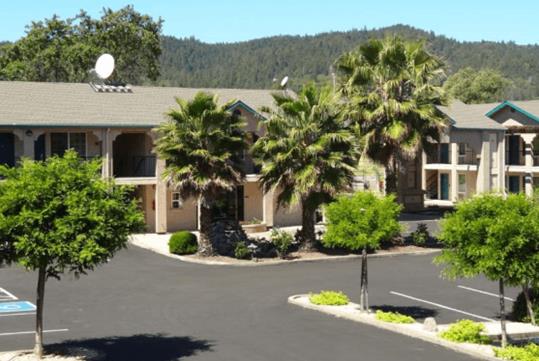 Exterior of the Cloverdale Wine Country Inn & Suites on a sunny day with several trees in the parking lot and in front of the building.