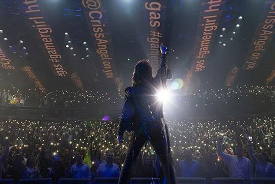 Criss Angel performing with his arm up on stage with a massive audience in front of him in Las Vegas.