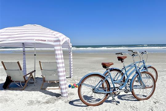 Two light blue and brown bikes parked next to a striped cabana with chairs under it on an empty beach.